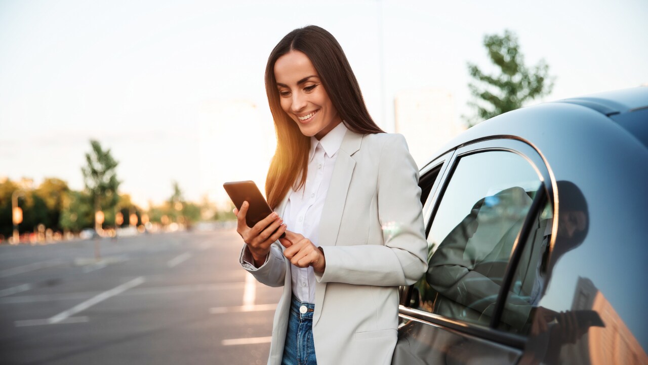 Mujer sonriendo mirando su teléfono mientras está de pie fuera de un auto.