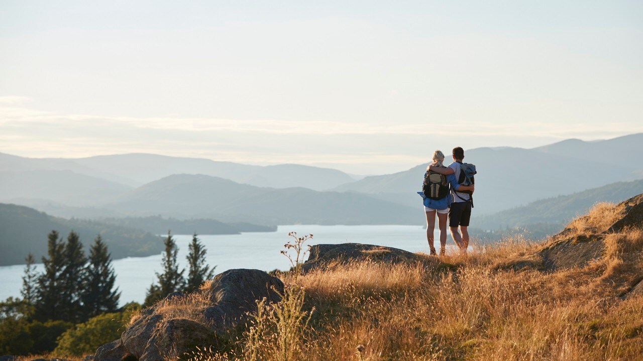 Rear View Of Couple Hugging On Top Of Hill On Hike Through Countryside In Lake District UK